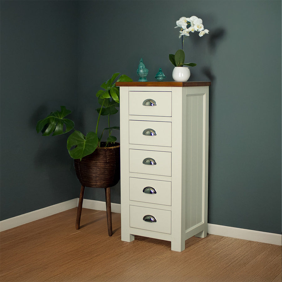 The front of the Alton 5 Drawer Lingerie Chest. There is a free standing potted plant next to it. There are two blue glass ornaments and a pot of white flowers on top.