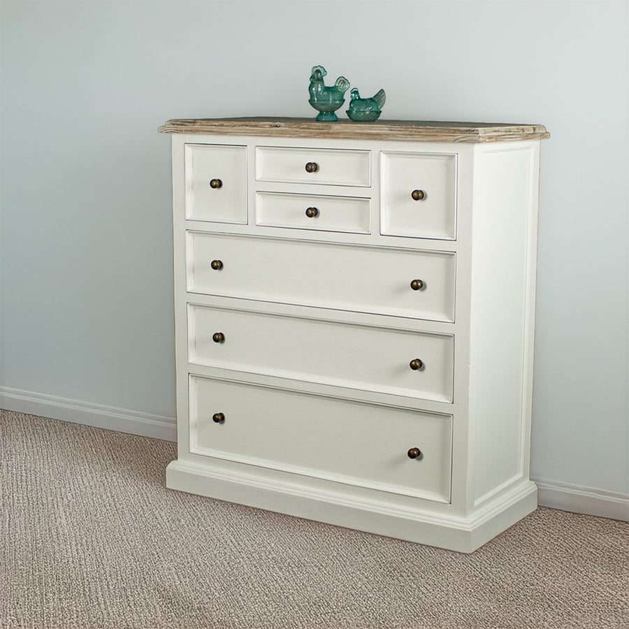 The front of the Biarritz 7 Drawer Tallboy / Chest of Drawer. There are two blue glass ornaments on top and a free standing potted plant next to it.