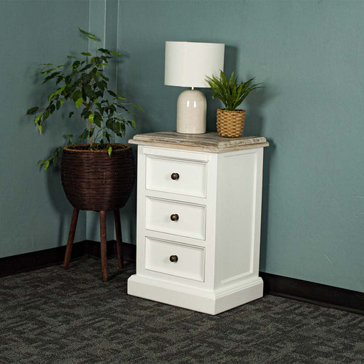 The front of the Biarritz 3 Drawer Bedside Cabinet with a lamp and potted plant on top. There is a potted plant next to the bedside table.