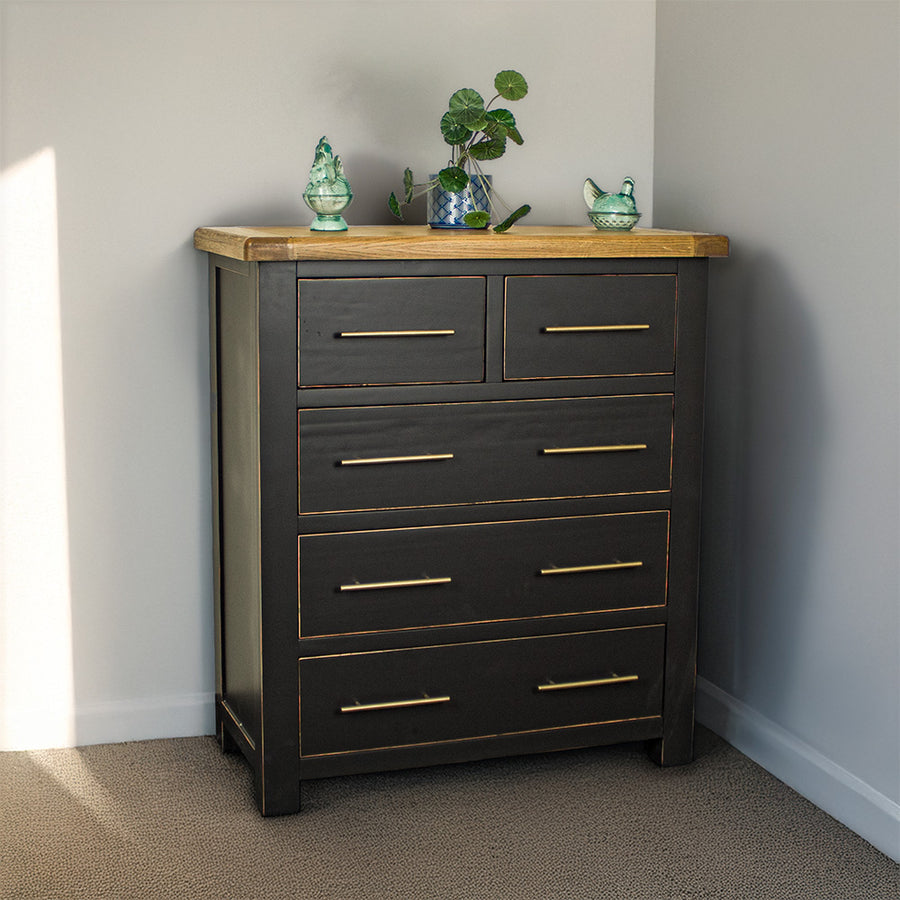 The front of the Cascais Oak Top Black Tallboy. There is a potted plant on top in the middle with two blue glass ornaments on either side.
