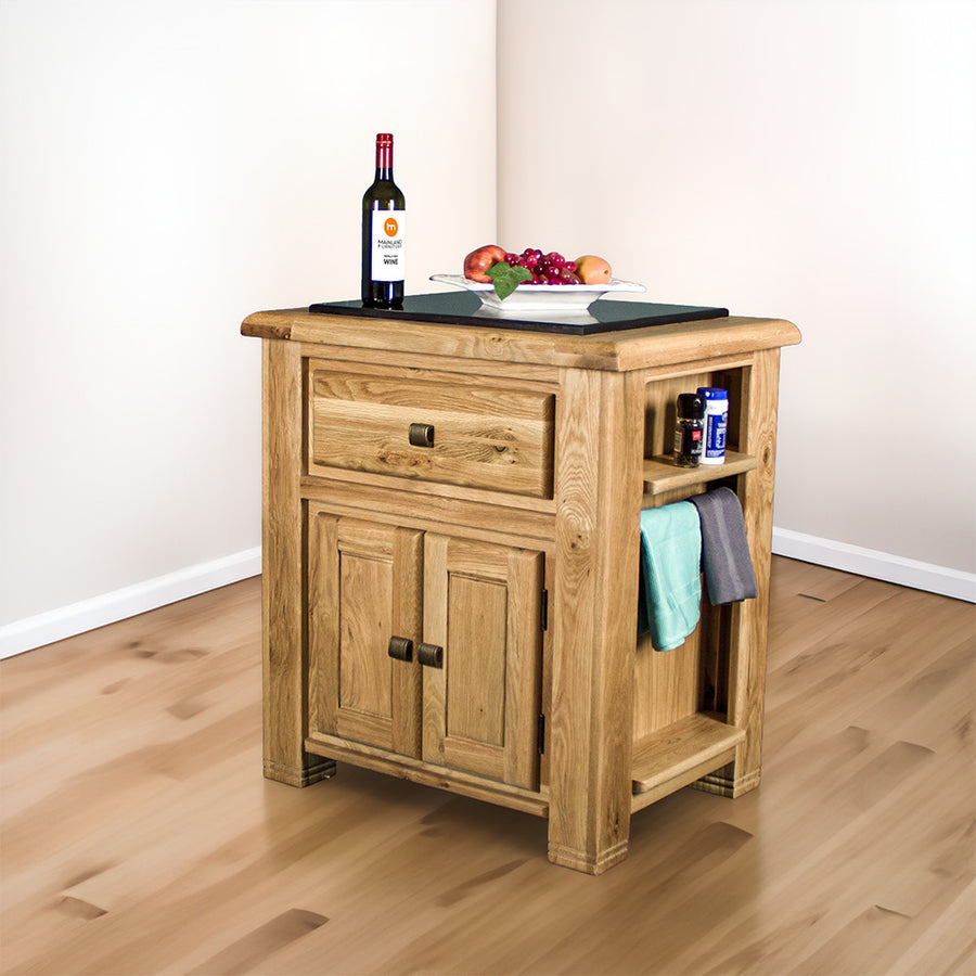 The front of the Danube Compact Granite Top Oak Kitchen Island, there is a fruit platter and a bottle of wine on the granite top, and two towels hanging on the side towel rack, with salt and pepper on the small shelf above it.
