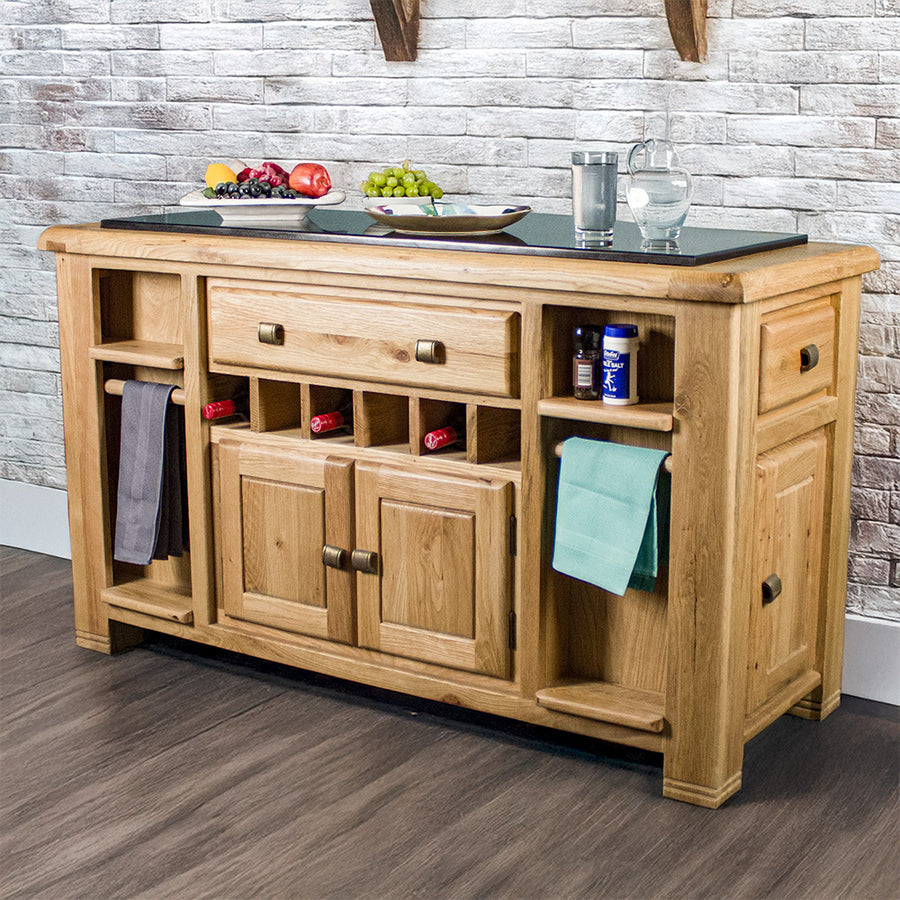 The front of the Danube Large Granite Top Oak Kitchen Island. There are two hand towels hanging on either side, three wine bottles in the wine rack on the middle, and a salt and pepper shaker on the small shelf in  the top right. There are two fruit platters on top, as well as a plate, a glass of water and a glass water pitcher.
