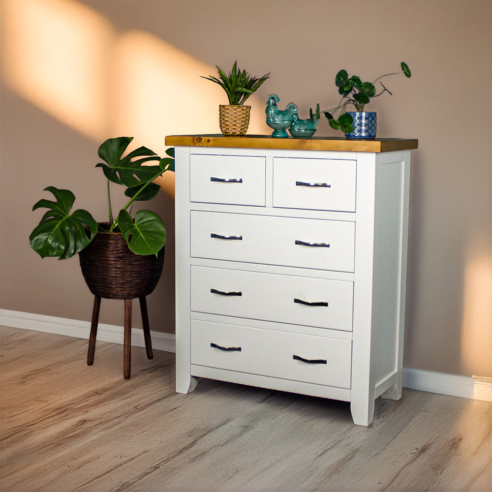 The front of the Felixstowe 5 Drawer Pine Tallboy. There are two potted plants on top with two blue glass ornaments in between. There is a free standing potted plant next to the tallboy.