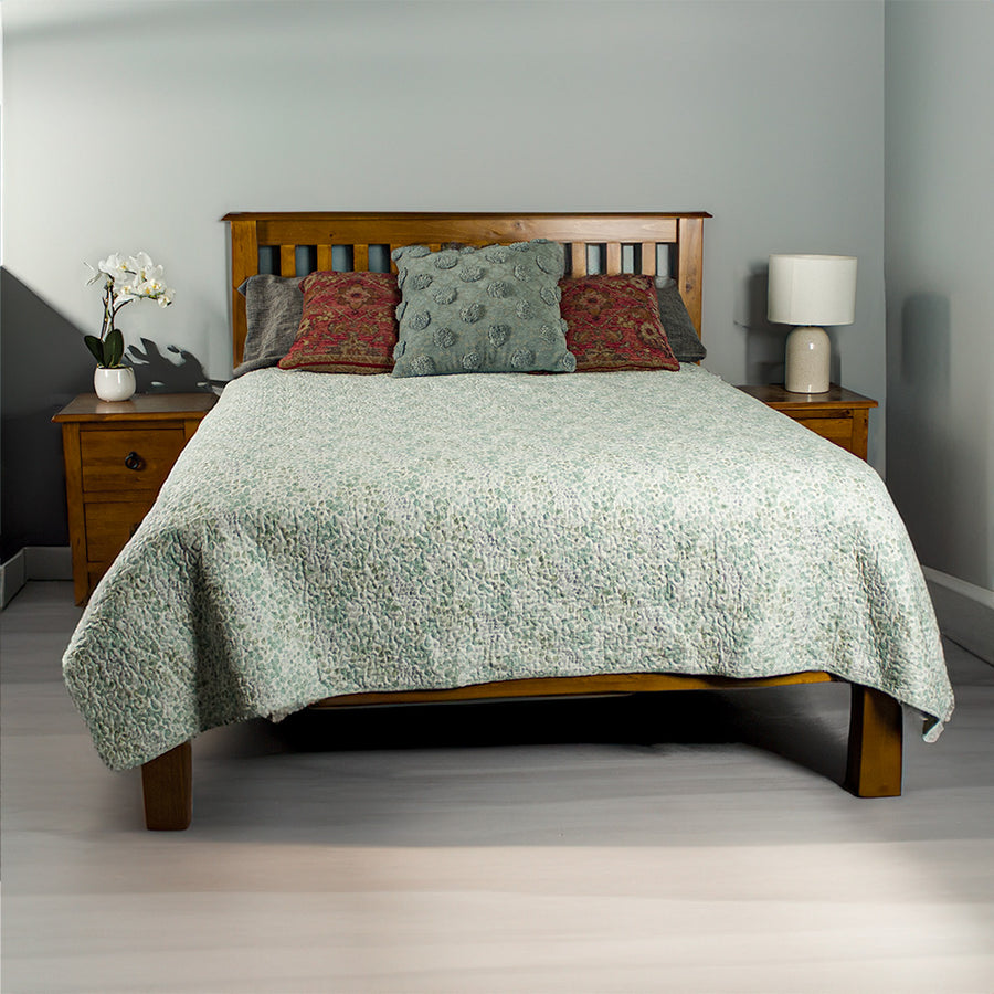 The front of the Trent Double Size NZ Pine Slat Bed Frame. There are two Trent Bedside Cabinets on either side. There is a pot of white flowers on the left side and a lamp on the right side. There are sheets and pillows on top of the bed frame.