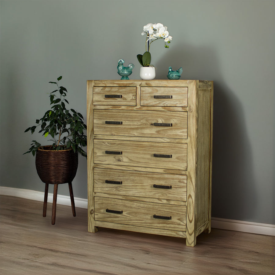 The front of the Vancouver 6 Drawer NZ Pine Tallboy. There are two blue glass ornaments on top with a small pot of white flowers in between. There is a free standing potted plant next to it.