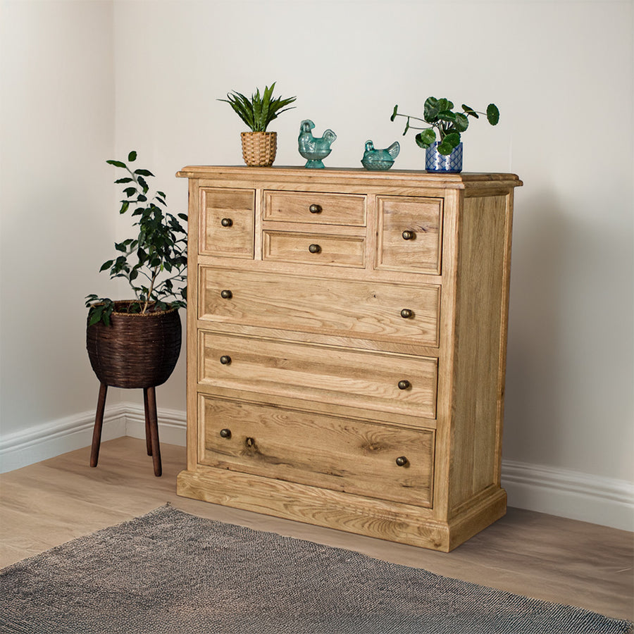 The front of the Versailles Oak 7 Drawer Chest. There are two potted plants on top, as well as two blue glass ornaments. There is a free standing potted plant next to it as well.