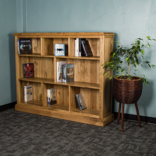 The front of the Versailles Outback Oak Three Level Bookcase. There are various books on each of the shelves, and a free standing potted plant next to the bookshelf.