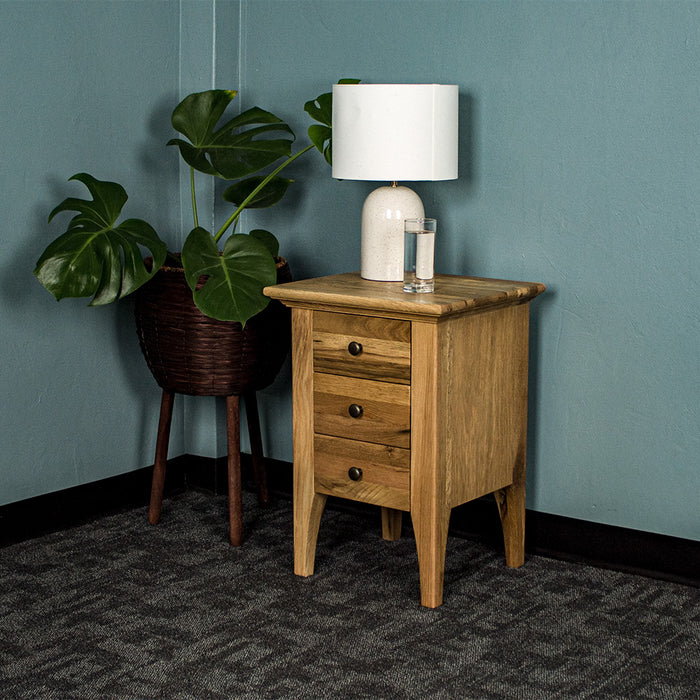 The front of the Beethoven Oak Bedside Table with 3 Drawers. There is a free standing potted plant next to it. There is a lamp and glass of water on top.