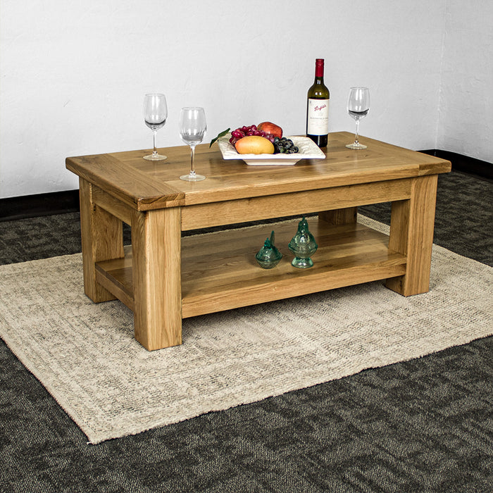 An overall of the Camden Oak Coffee Table. There are three wine glasses, a bottle of wine and a fruit platter on top. There are two blue glass ornaments on the lower shelf.