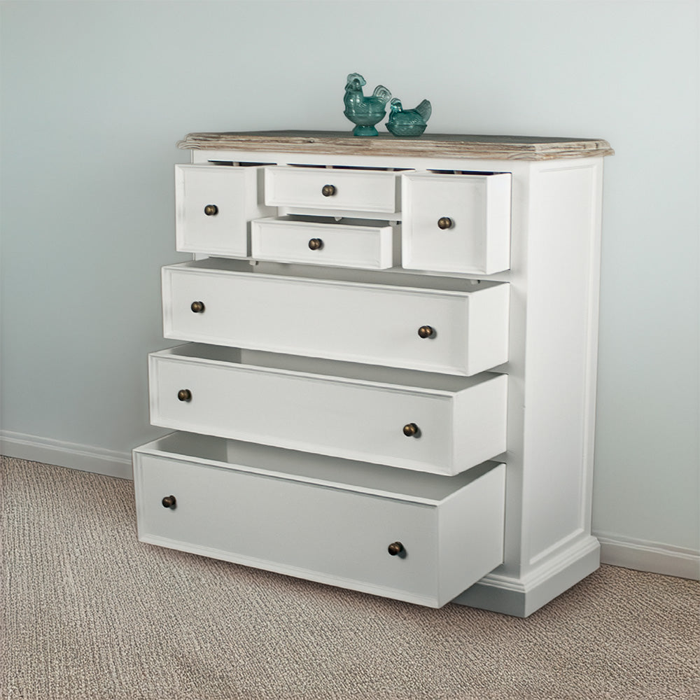 The front of the Biarritz 7 Drawer Tallboy / Chest of Drawer with its drawers open. There are two blue glass ornaments on top and a free standing potted plant next to it.