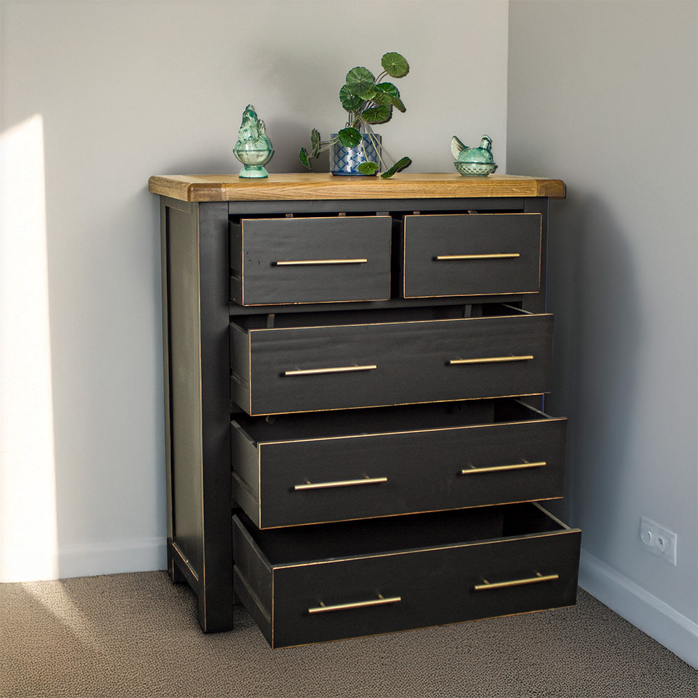 The front of the Cascais Oak Top Black Tallboy with its drawers open. There is a potted plant on top in the middle with two blue glass ornaments on either side.