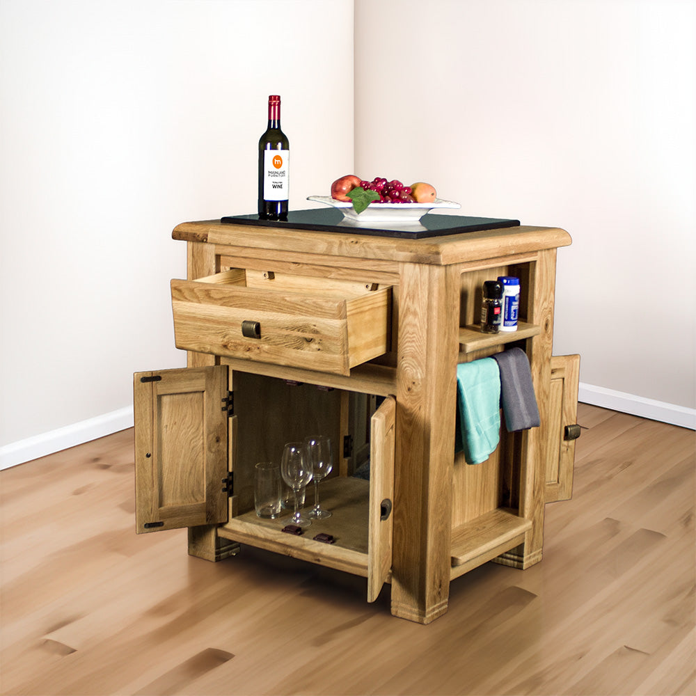 The front of the Danube Compact Granite Top Oak Kitchen Island with its doors and drawers open, there is a fruit platter and a bottle of wine on the granite top, and two towels hanging on the side towel rack, with salt and pepper on the small shelf above it. There are two sets of glasses and wine glasses in the middle shelf/opening.