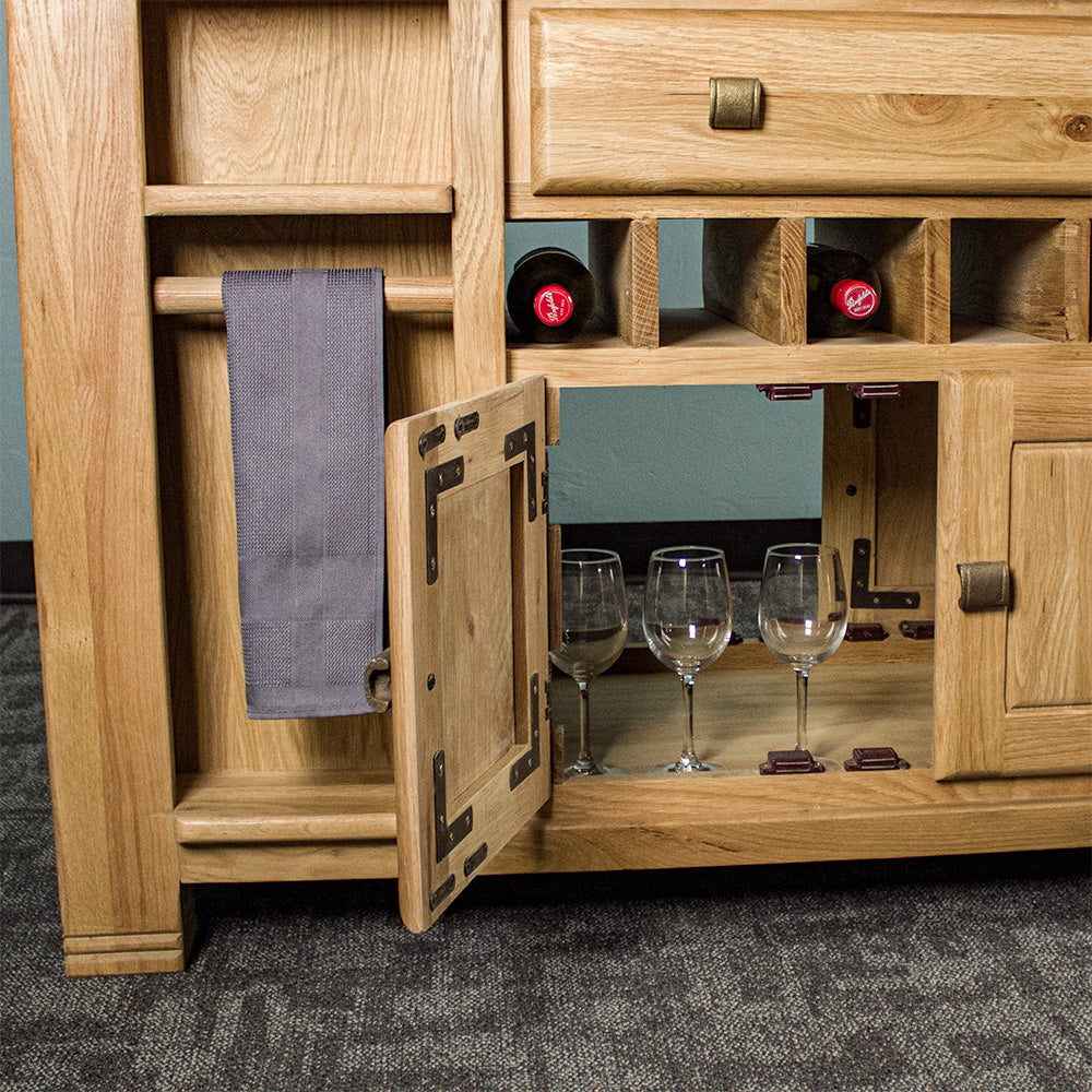 A closer view of the Danube Large Granite Top Oak Kitchen Island's open ended lower shelf. There are three wine glasses inside. There are three wine bottles on the small wine rack above and a hand towel hanging on the towel rail to the left.