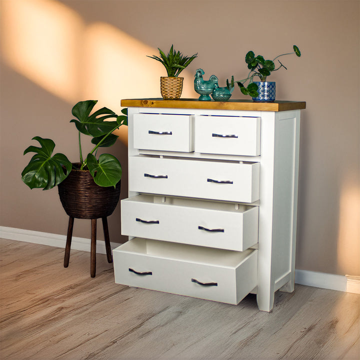 The front of the Felixstowe 5 Drawer Pine Tallboy, with its drawers open. There are two potted plants on top with two blue glass ornaments in between. There is a free standing potted plant next to the tallboy.