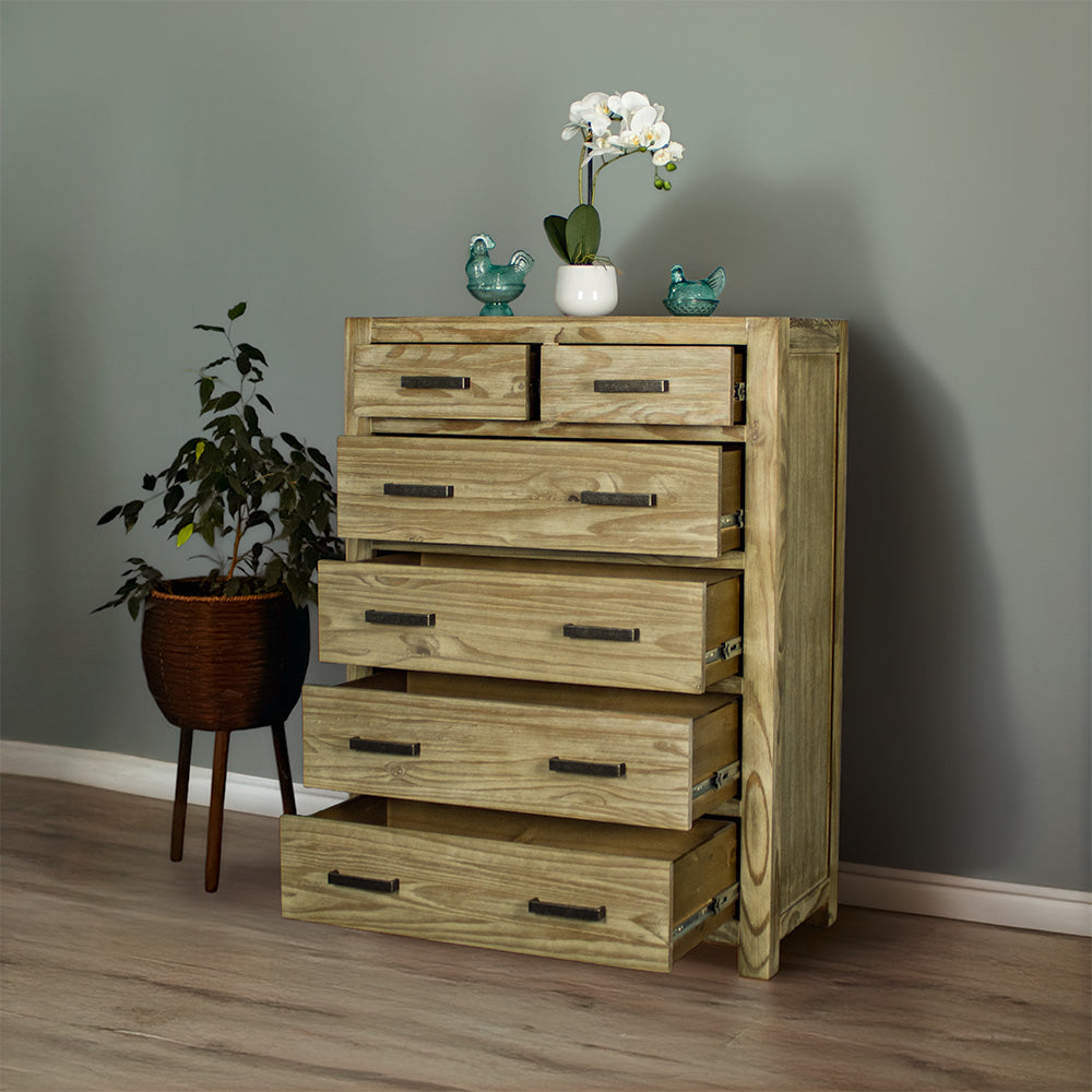 The front of the Vancouver 6 Drawer NZ Pine Tallboy with its drawers open. There are two blue glass ornaments on top with a small pot of white flowers in between. There is a free standing potted plant next to the unit.