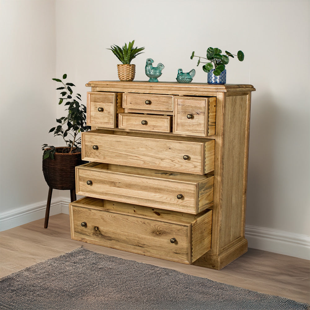 The front of the Versailles Oak 7 Drawer Chest with its drawers open. There are two potted plants and two blue glass ornaments on top. There is a free standing potted plant next to it.