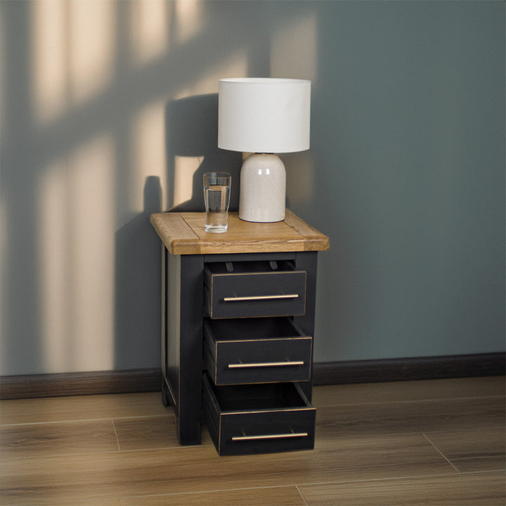 The front of the Cascais 3 Drawer Black Bedside Table with its drawers open. There is a glass of water and a lamp on top.
