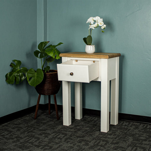 The front of the Loire Oak Console Table with its drawer open. There is a pot of white flowers on top and a free standing potted plant to the right.