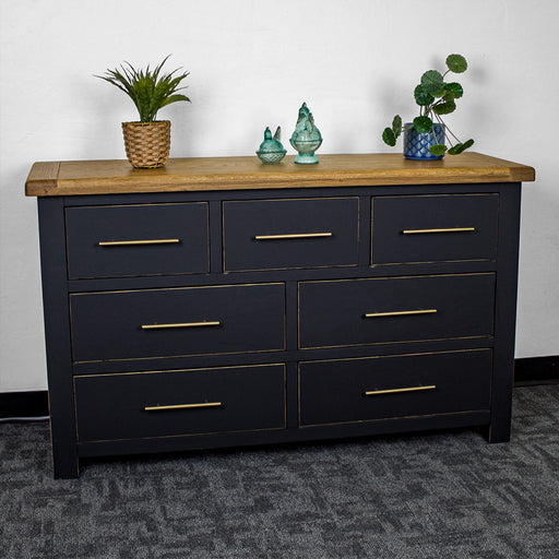 The front of the Cascais Oak Top 7 Drawer Lowboy (Black). There are two blue glass ornaments on top in the middle and two potted plants on either side.