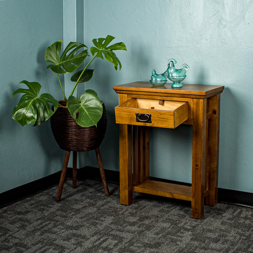 The front of the Montreal Small Pine Hall Table with its drawer open. There are two blue glass ornaments on top with a free standing potted plant next to it.