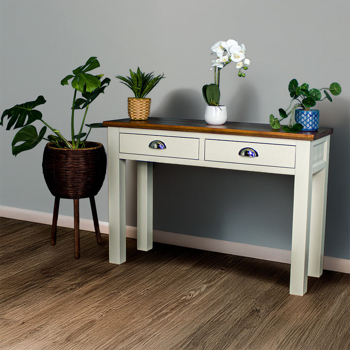 White Alton 2 Drawer Pine Hall Table with a rimu stained top. There are two potted plants on the top and a bouquet of flowers in the middle.