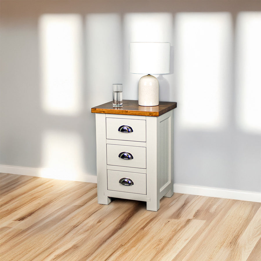 The Alton 3 Drawer Pine Bedside Cabinet with a Rimu stained top. There is a lamp on top and a free standing potted plant next to it.