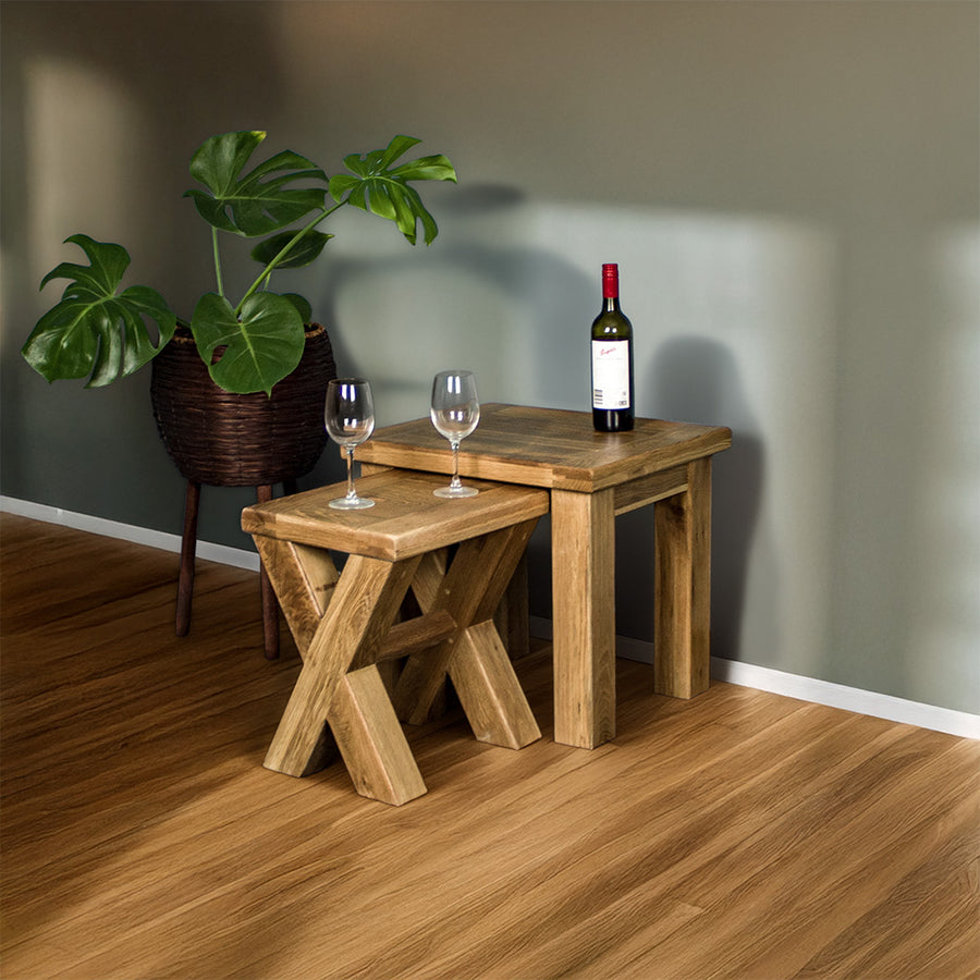 An overall view of the Amstel Oak Nesting Tables. The smaller table is out from underneath the top one. There is a free standing potted plant next to the tables. There are two wine glasses on top of the smaller table and a bottle of wine on the larger table.