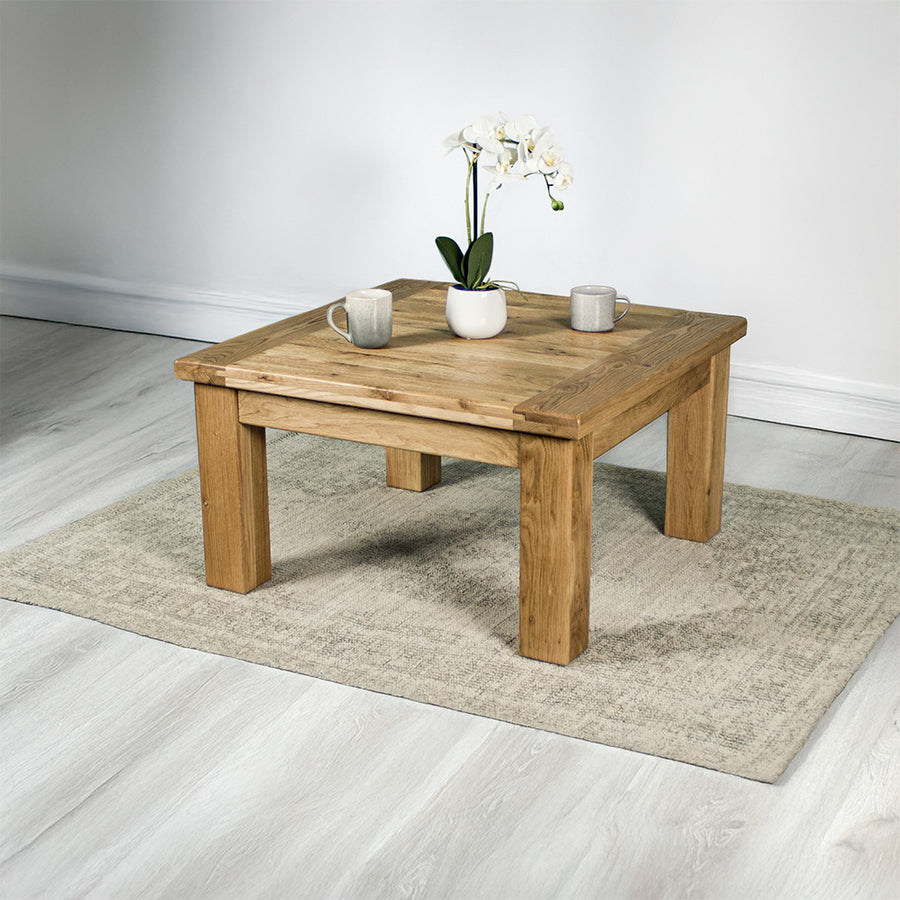The front of the Amstel Square Oak Coffee Table. There is a pot of white flowers and two coffee mugs on top. The coffee table is on a white rug.
