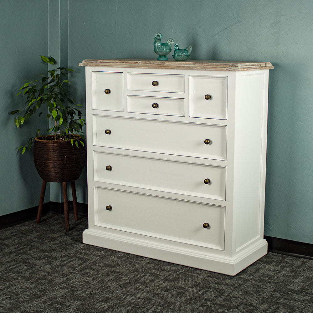 The front of the Biarritz 7 Drawer Tallboy / Chest of Drawer. There are two blue glass ornaments on top and a free standing potted plant next to it.