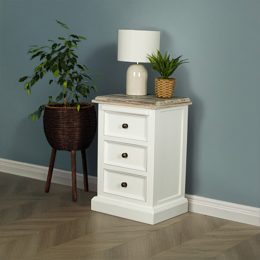 The front of the Biarritz 3 Drawer Bedside Cabinet with a lamp and potted plant on top. There is a potted plant next to the bedside table.