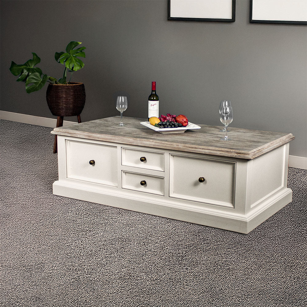 An overall view of the Biarritz 4 Drawer Extra-Large Coffee Table. There are three wine glasses, a bottle and a platter of fruit on top. There is a free standing potted plant in the background.