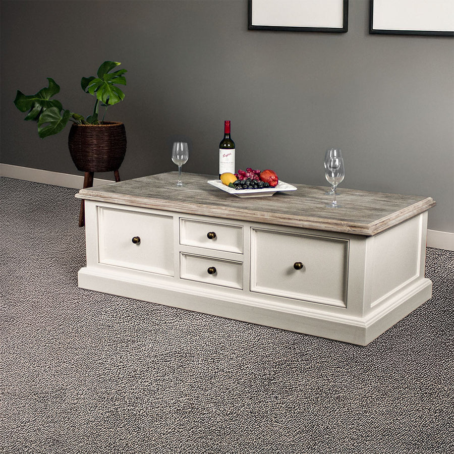 An overall view of the Biarritz 4 Drawer Extra-Large Coffee Table. There are three wine glasses, a bottle and a platter of fruit on top. There is a free standing potted plant in the background.