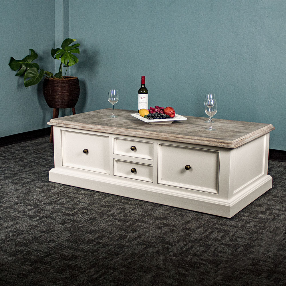 An overall view of the Biarritz 4 Drawer Extra-Large Coffee Table. There are three wine glasses, a bottle and a platter of fruit on top. There is a free standing potted plant in the background.