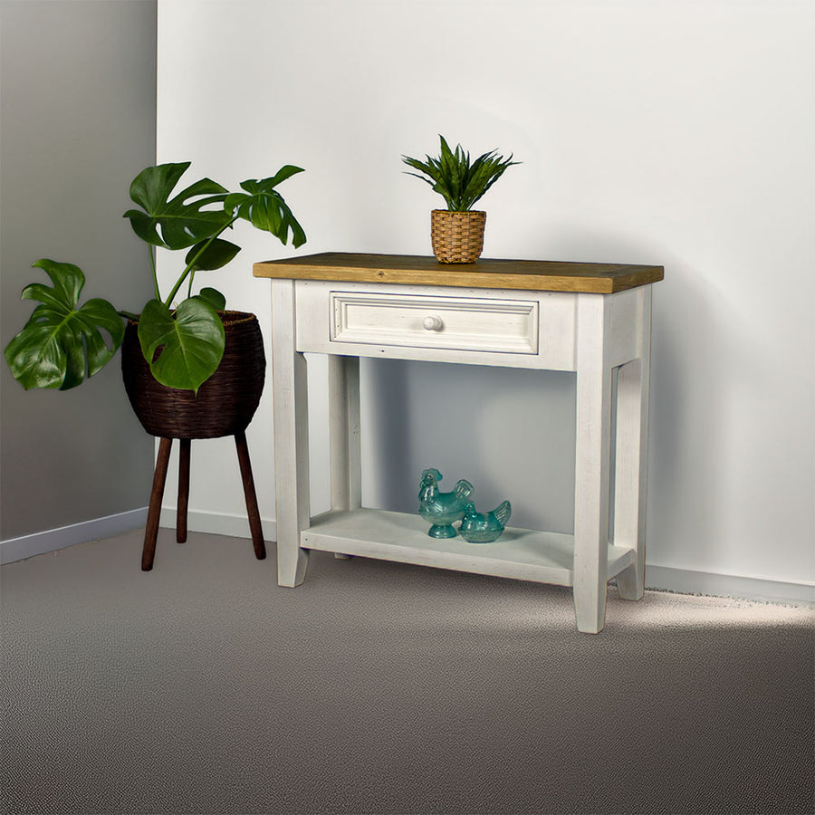 Front view of the Byron Recycled Pine Hall Table. There is a standing potted plant to the side, a small potted plant on top and two blue glass ornaments on the lower shelf.