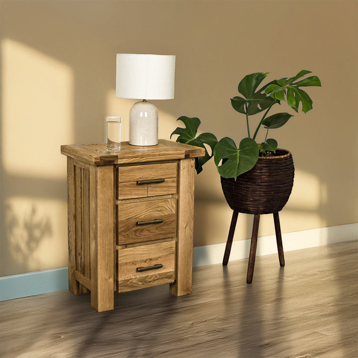An overall view of the Camden 3 Drawer White Oak Bedside Table. There is a glass of water and a lamp on top. There is a free standing potted plant next to the bedside table.
