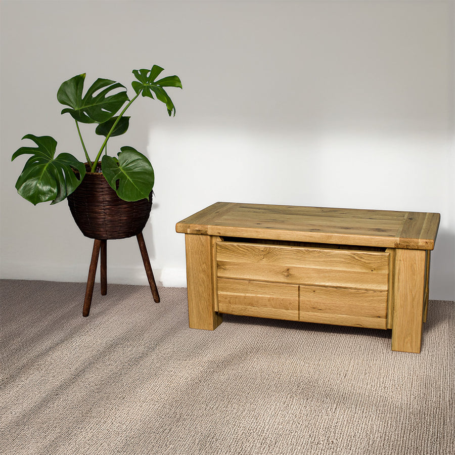 An overall view of the Camden Oak Blanket Box. There is a free standing potted plant next to it.