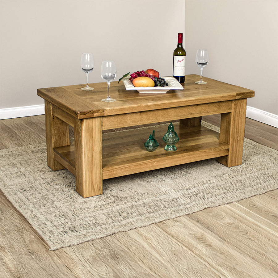 An overall of the Camden Oak Coffee Table. There are three wine glasses, a bottle of wine and a fruit platter on top. There are two blue glass ornaments on the lower shelf.