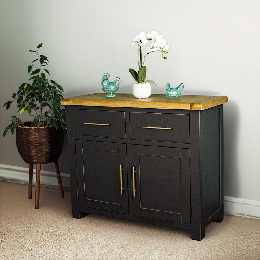 The front of the Cascais Oak-Top Two Drawer Buffet. There are two blue glass ornaments on top with a pot of white flowers in between. There is a free standing potted plant next to the sideboard.