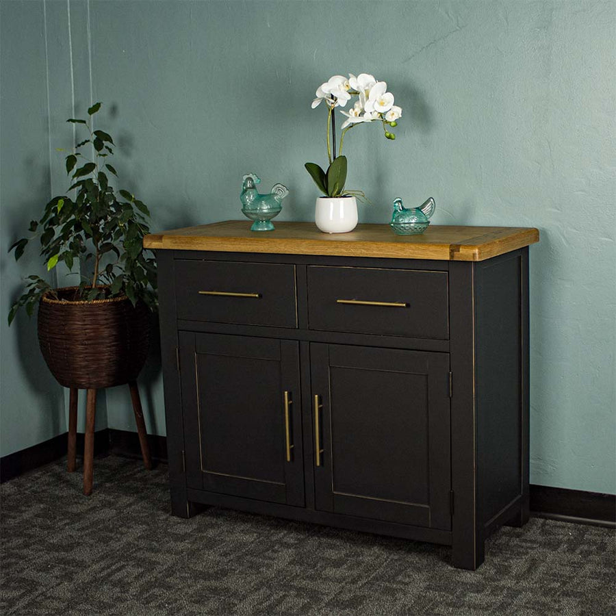 The front of the Cascais Oak-Top Two Drawer Buffet. There are two blue glass ornaments on top with a pot of white flowers in between. There is a free standing potted plant next to the sideboard.