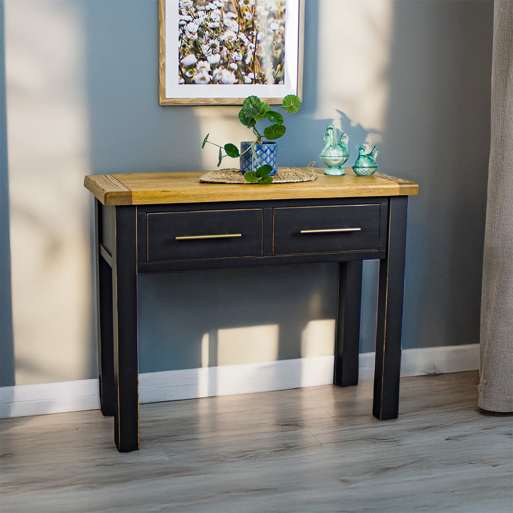 The front of the Cascais 2-Drawer Hall Table. There is a potted plant on top of a rattan leaf, in the middle on the top. There are two blue glass ornaments on top. There is a picture of flowers on the wall.
