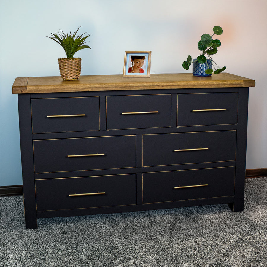 The front of the Cascais Oak Top 7 Drawer Lowboy (Black). There are two blue glass ornaments on top in the middle and two potted plants on either side.