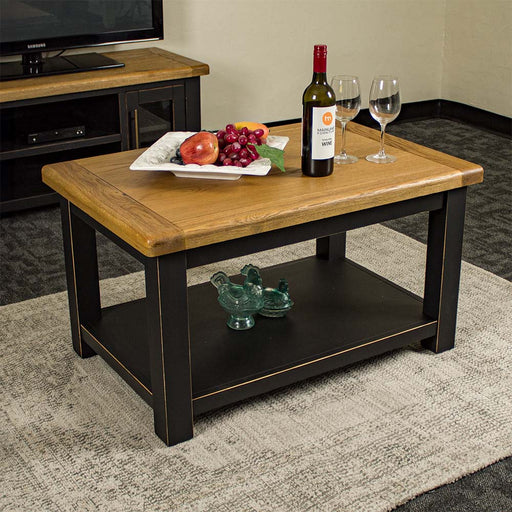 The front of the Cascais Oak-Top Coffee Table. There is a platter of fruit, a bottle of wine and two wine glasses on top. There are two blue glass ornaments on the shelf below.