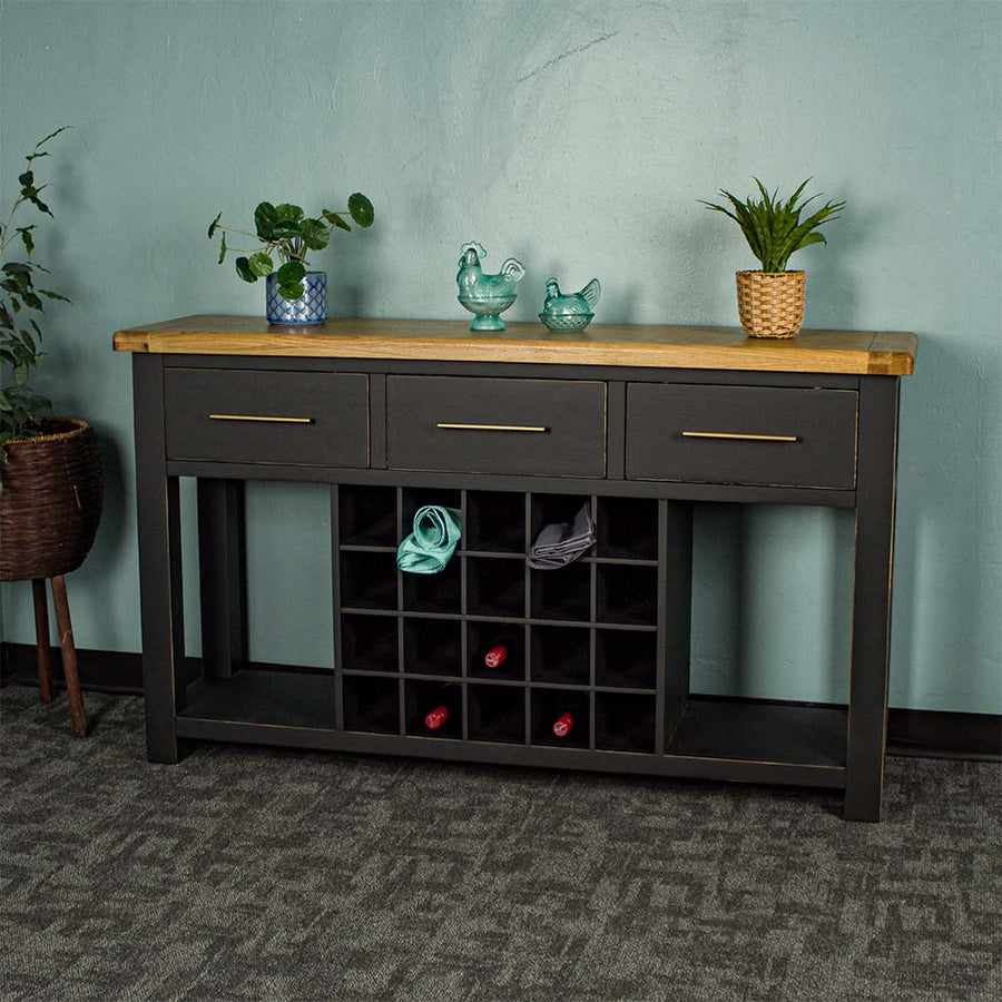 The front of the Cascais Large Buffet with Wine Rack. There is a tall potted plant next to the unit. There are two small potted plants on top in between two blue glass ornaments. There are three wine bottles in the wine rack and two towels in the top.