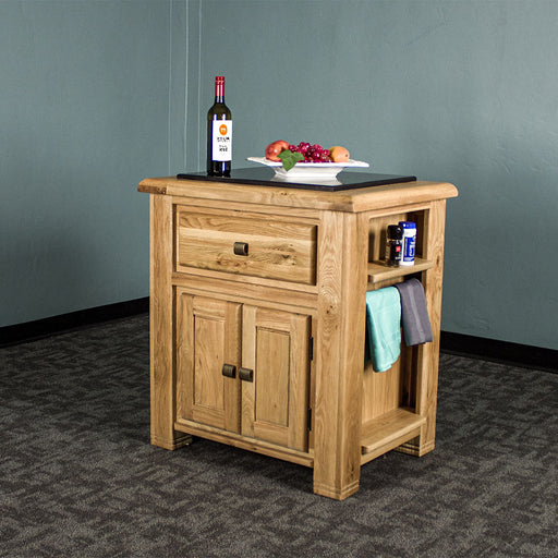 The front of the Danube Compact Granite Top Oak Kitchen Island, there is a fruit platter and a bottle of wine on the granite top, and two towels hanging on the side towel rack, with salt and pepper on the small shelf above it.
