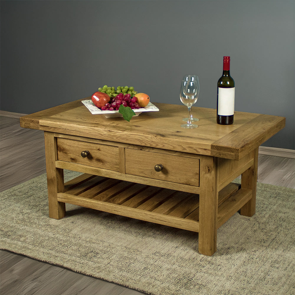 The front of the Farmhouse Coffee Table, which is sitting on a gray rug. There is a free standing potted plant in the back. There is a fruit platter, two wine glasses and a wine bottle on top.