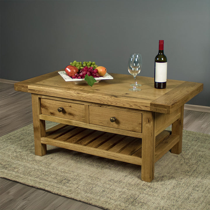 The front of the Farmhouse Coffee Table, which is sitting on a gray rug. There is a free standing potted plant in the back. There is a fruit platter, two wine glasses and a wine bottle on top.