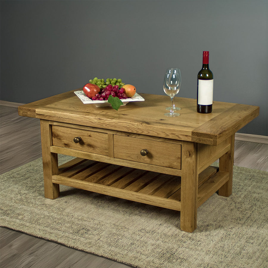 The front of the Farmhouse Coffee Table, which is sitting on a gray rug. There is a free standing potted plant in the back. There is a fruit platter, two wine glasses and a wine bottle on top.