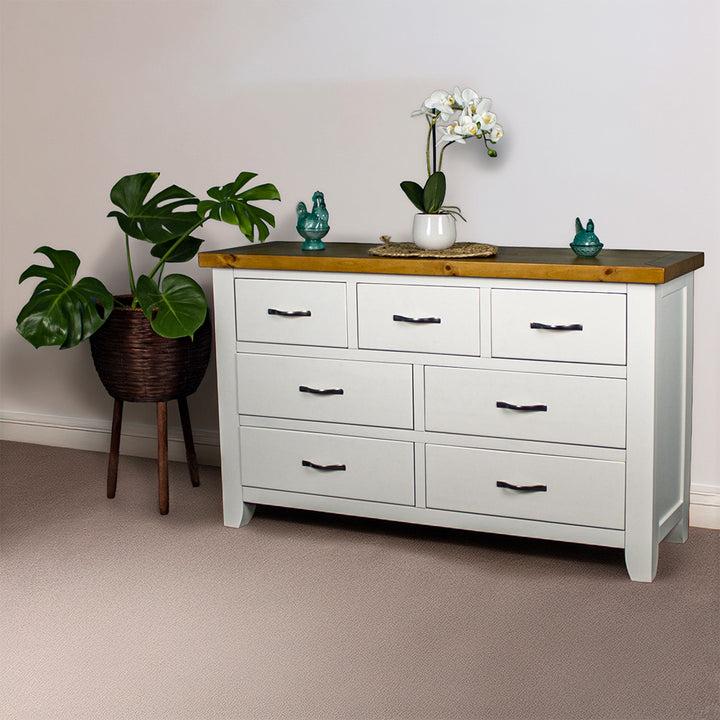 The front of the Felixstowe 7 Drawer Pine Lowboy. There are two blue glass ornaments on top with a small pot of white flowers on a rattan leaf in the middle of them. There is a free standing potted plant next to the lowboy.