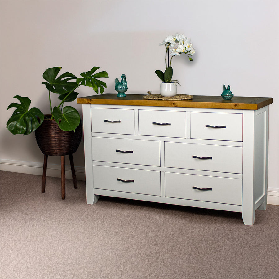 The front of the Felixstowe 7 Drawer Pine Lowboy. There are two blue glass ornaments on top with a small pot of white flowers on a rattan leaf in the middle of them. There is a free standing potted plant next to the lowboy.