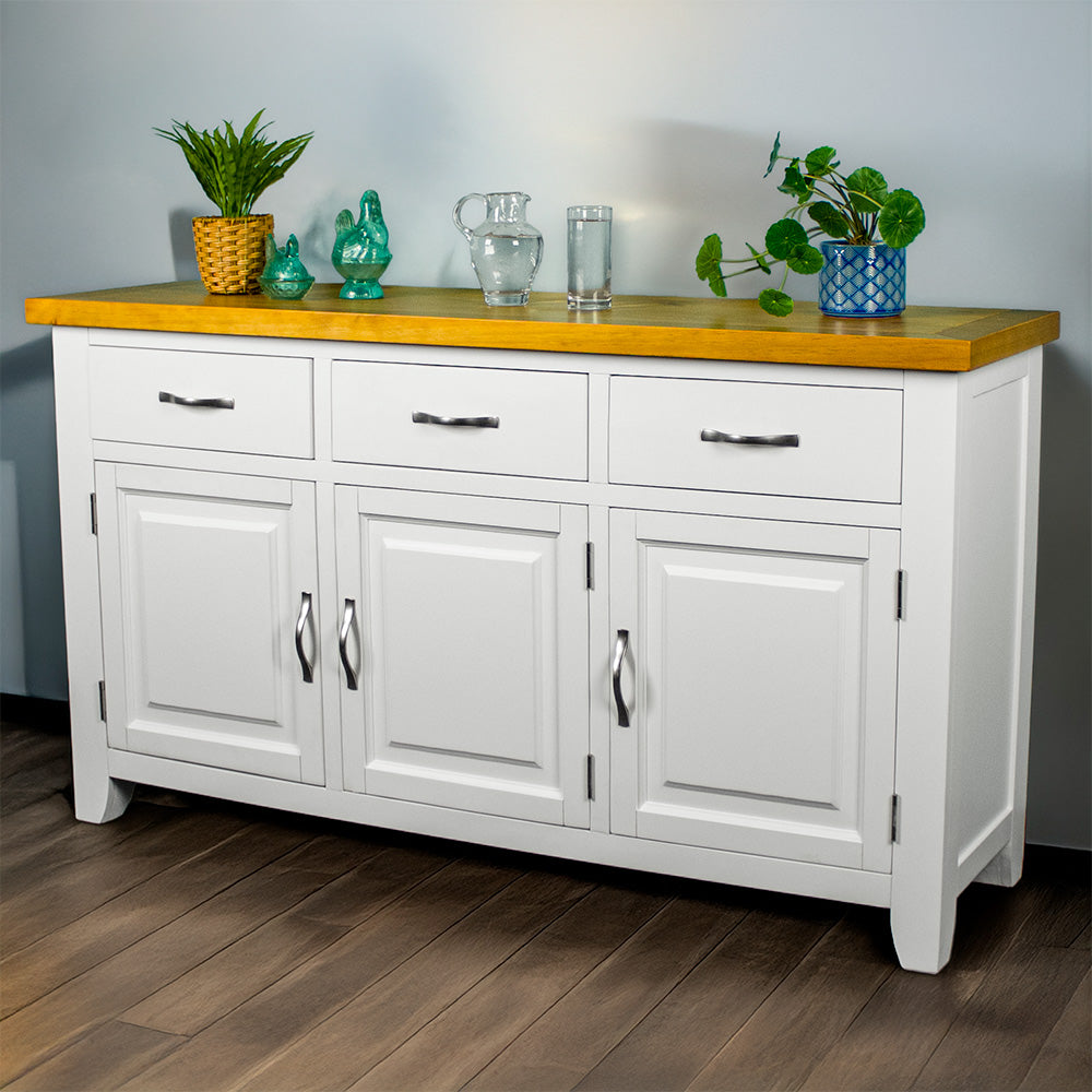 The front of the Felixstowe Large Pine White Buffet. From left to right there are: a potted plant, two blue glass ornaments, a water pitcher, a glass of water and a potted plant on top of the sideboard.